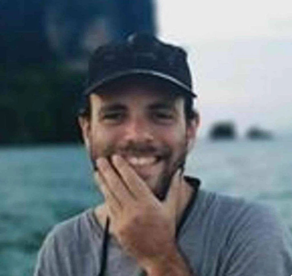 a man with a hat smiling in front of a body of water.