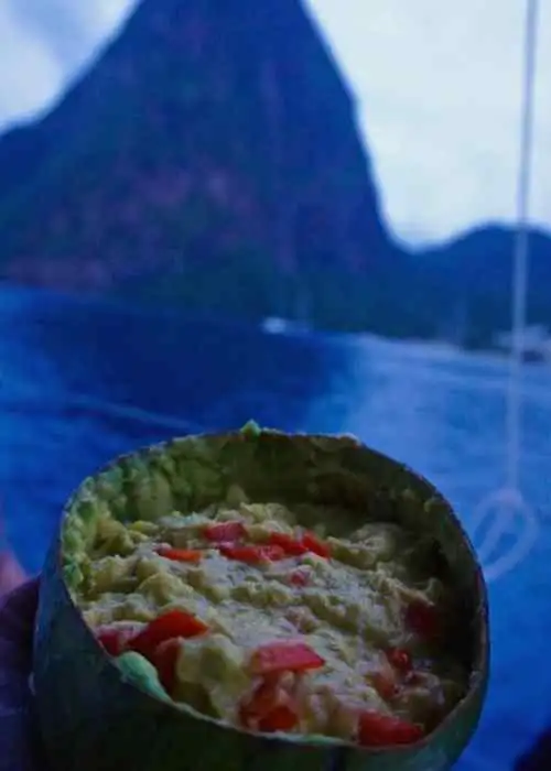 An offshore sailing crew member holds a bowl of food on a boat with majestic mountains in the background.