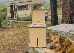 A dog sits next to a toilet box in front of an RV, highlighting the environmentally friendly choice of a compost toilet.