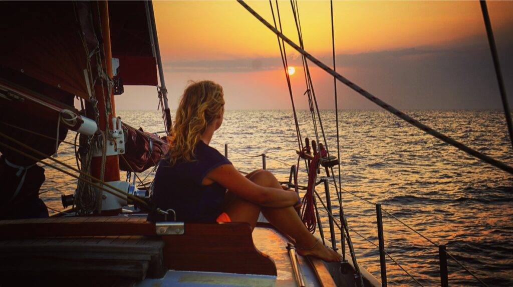 A woman is sailing on the deck of a sailboat at sunset.
