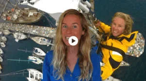 A woman in a yellow jacket is standing next to a group of sailboats, surrounded by a sailing crew.
