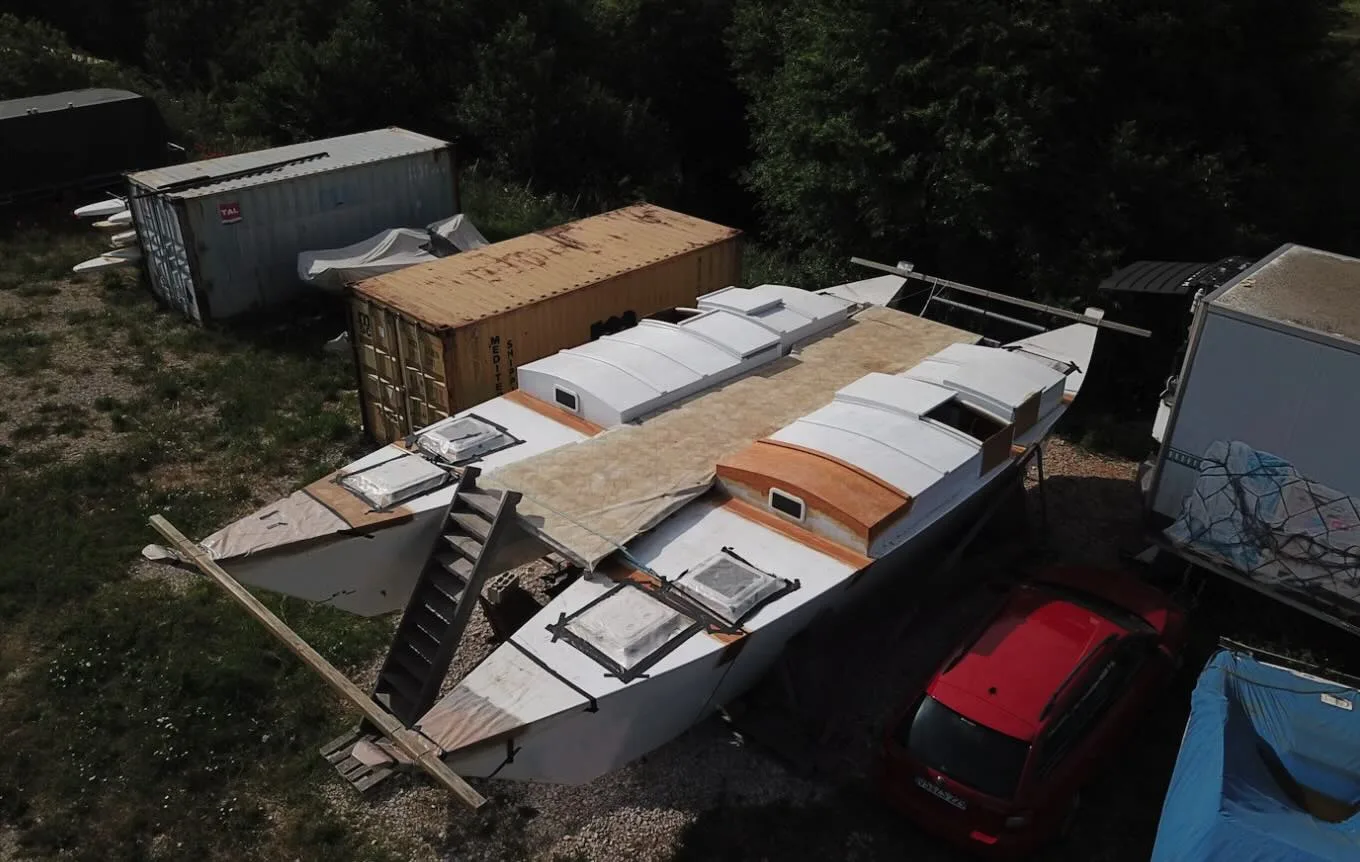 A catamaran is under construction/repair outdoors. It's surrounded by shipping containers, one of which is yellow, and a red car, signaling active sailboat travel and crewing opportunities.