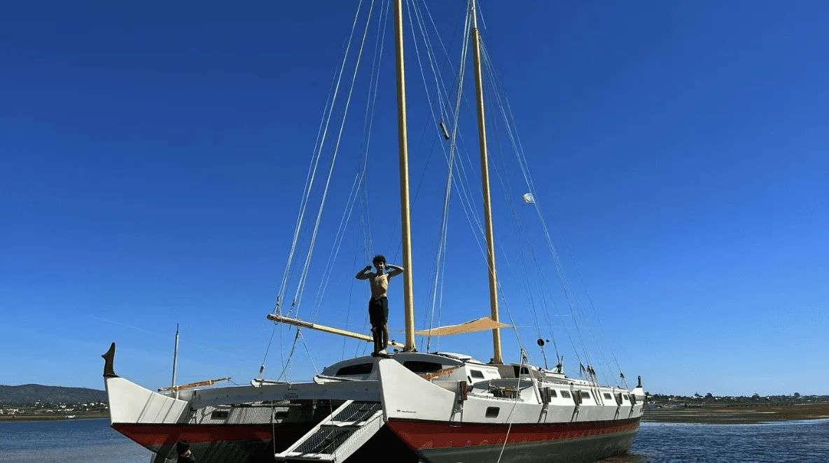 A person stands on the deck of a large sailboat with tall masts, docked in a calm body of water under a clear blue sky, dreaming of sailboat travel and crewing opportunities that lie ahead.