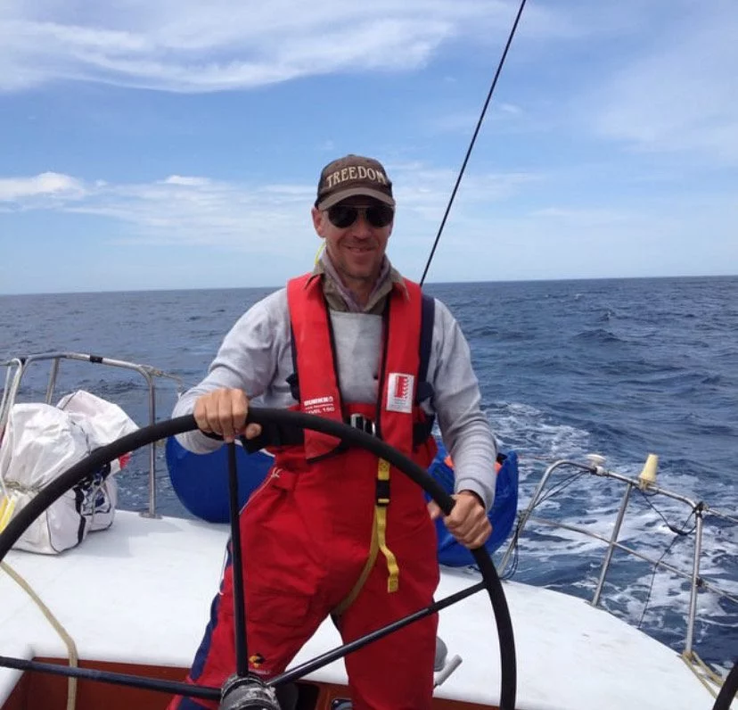 A person wearing a red life jacket and a cap, resembling an experienced skipper, steers a sailboat on open water under a cloudy sky. Blair seems at home among the Ocean Nomads.