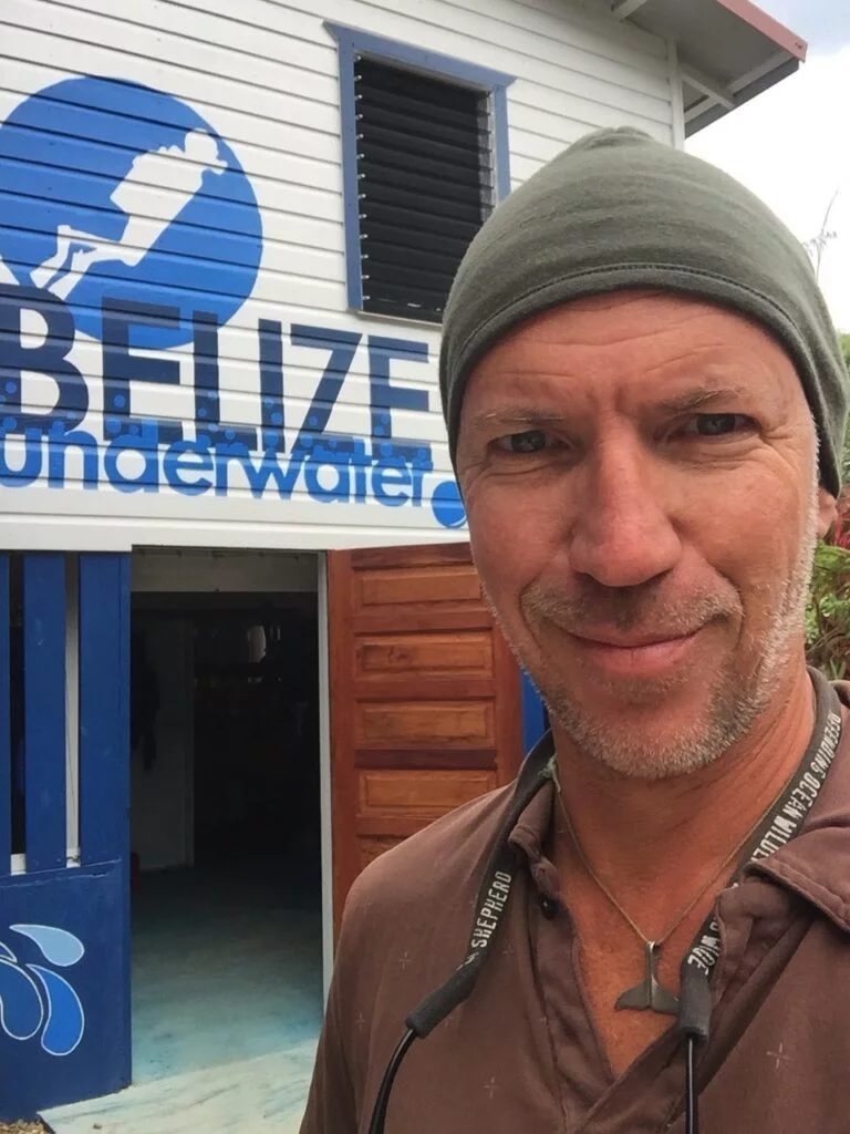 A person wearing a beanie and a lanyard takes a selfie in front of the "Belize Underwater" sign, looking every bit like an Ocean Nomad on an adventure.