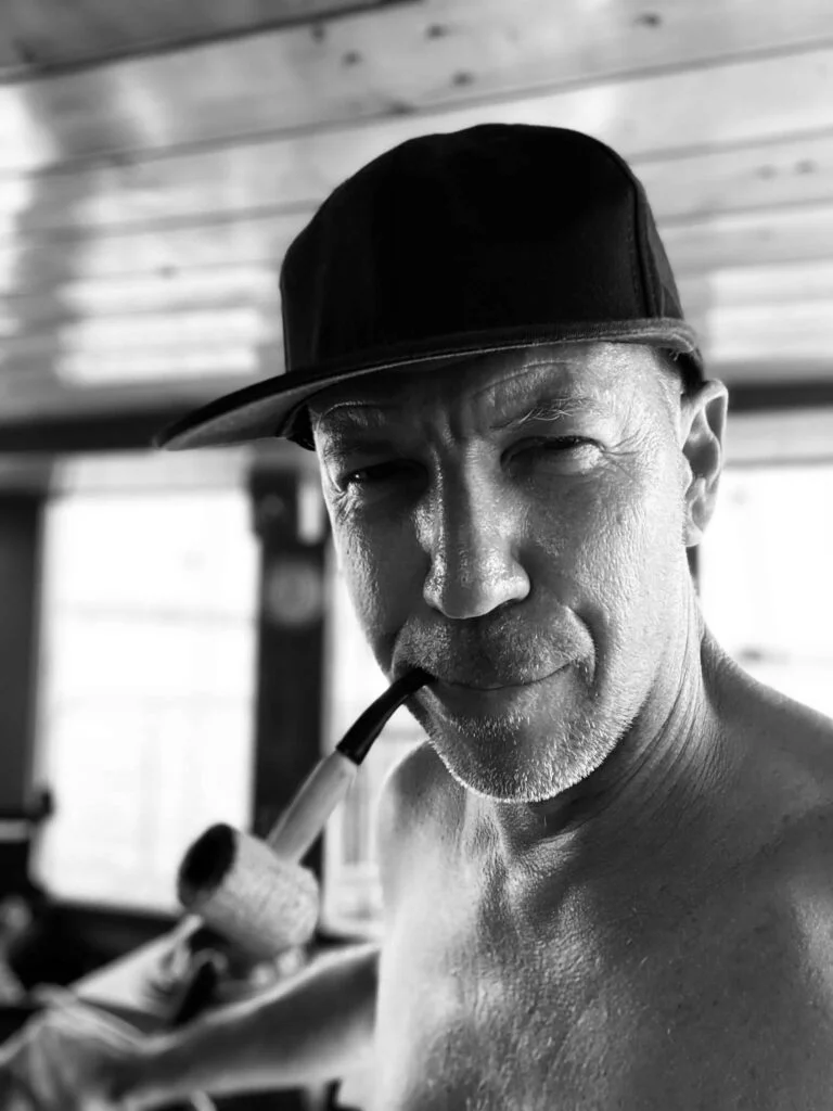 Black and white image of Blair, a shirtless man wearing a cap, smoking a pipe, and looking at the camera indoors. Skipper of the Ocean Nomads.