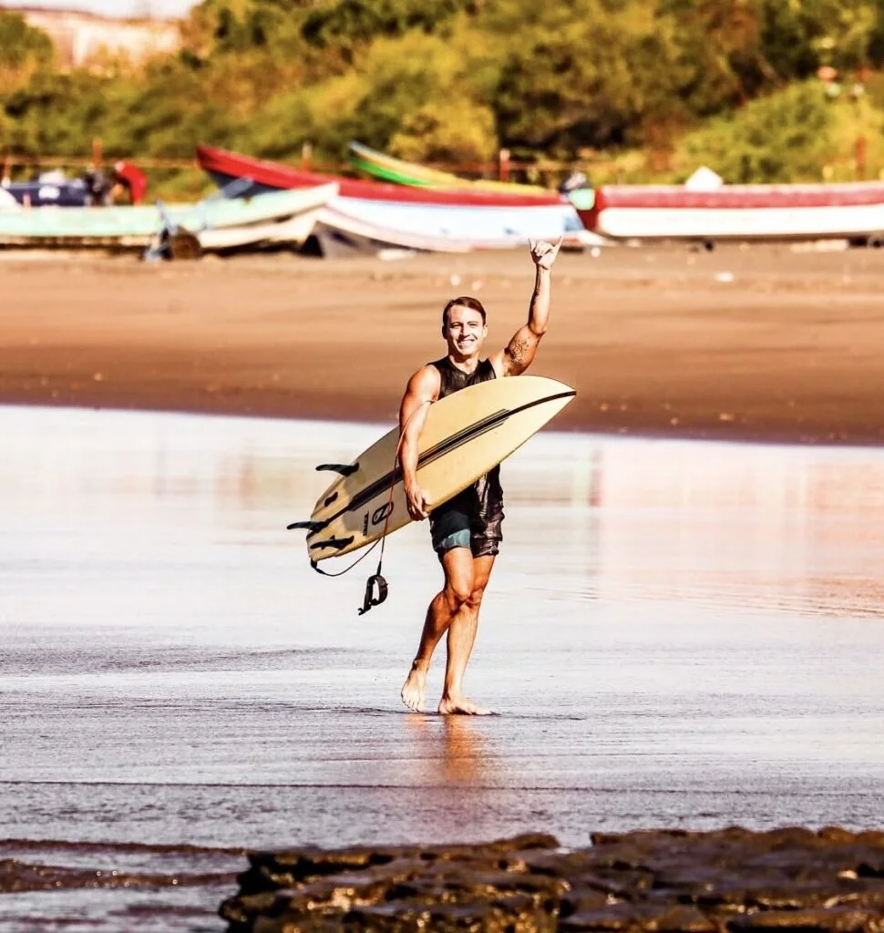 A surfer holding a surfboard waves while walking on a beach with boats in the background.