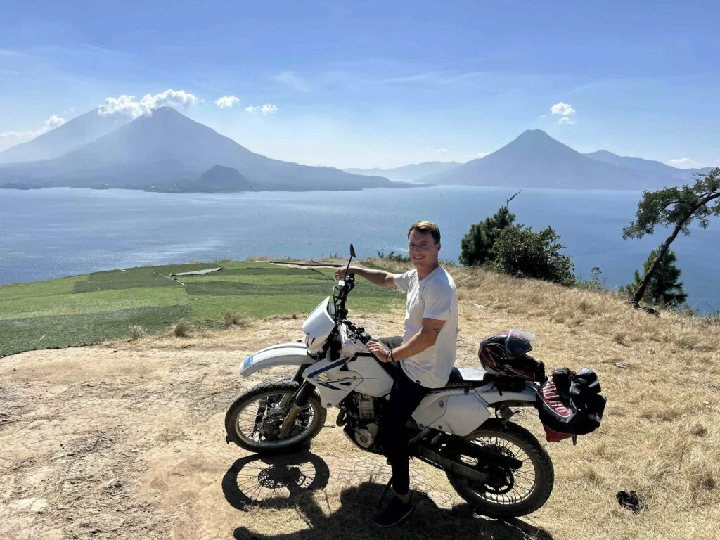 A world sailor sits on a motorcycle near a grassy cliff with a scenic view of a lake and distant mountains in the background.