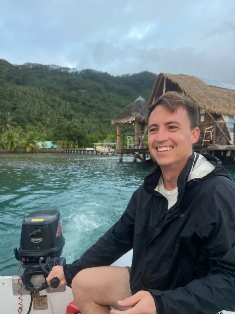A world sailor in a black jacket smiles while operating a boat. In the background, there are overwater bungalows, greenery, and a mountainous landscape.