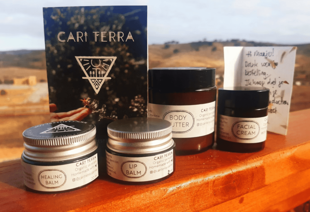 Four jars labeled healing balm, lip balm, body butter, and facial cream from CARI TERRA are displayed on a wooden surface—a perfect pick for Eco Friendly Gifts and thoughtful Nature Connected Nomads. A postcard and handwritten note add a personal touch.
