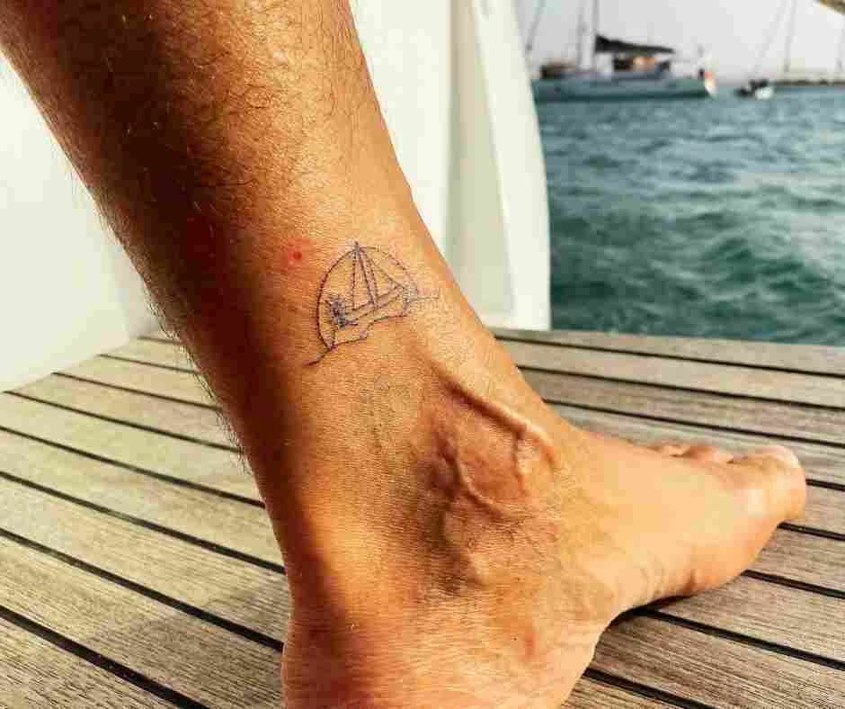 A person's ankle adorned with a small sailboat tattoo, poised on a wooden deck with the vast ocean stretching toward Cape Verde in the background.