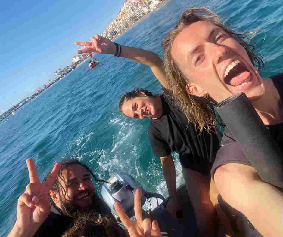 Three people smiling and making peace signs on a boat in the ocean, with a stunning cityscape in the background, set against the vibrant backdrop of Cape Verde.