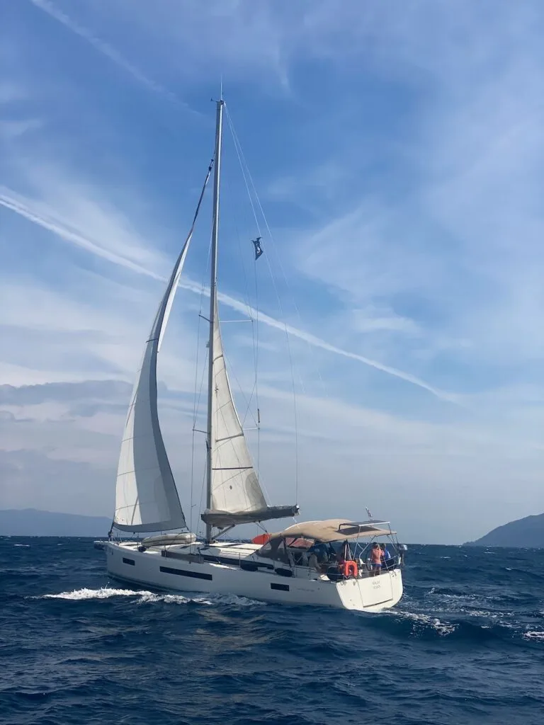 A sailboat with white sails navigates through a slightly choppy sea under a blue sky with wispy clouds, engaging in offshore sailing. Several people are on the boat, possibly in sail training, and there is a distant landmass hinting at the Canary Islands on the horizon.