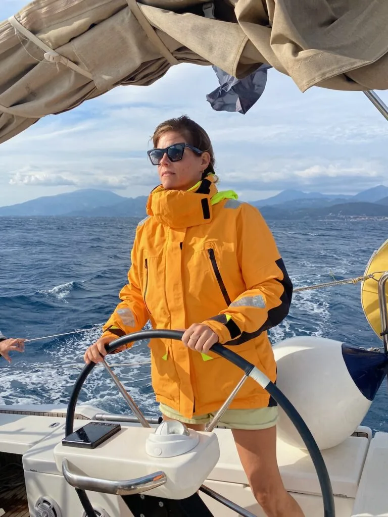 Wearing a yellow jacket and sunglasses, a person expertly navigates the sailboat through the deep blue waters near the Canary Islands, with majestic mountains forming a stunning backdrop. It's a perfect example of skillful offshore sailing.