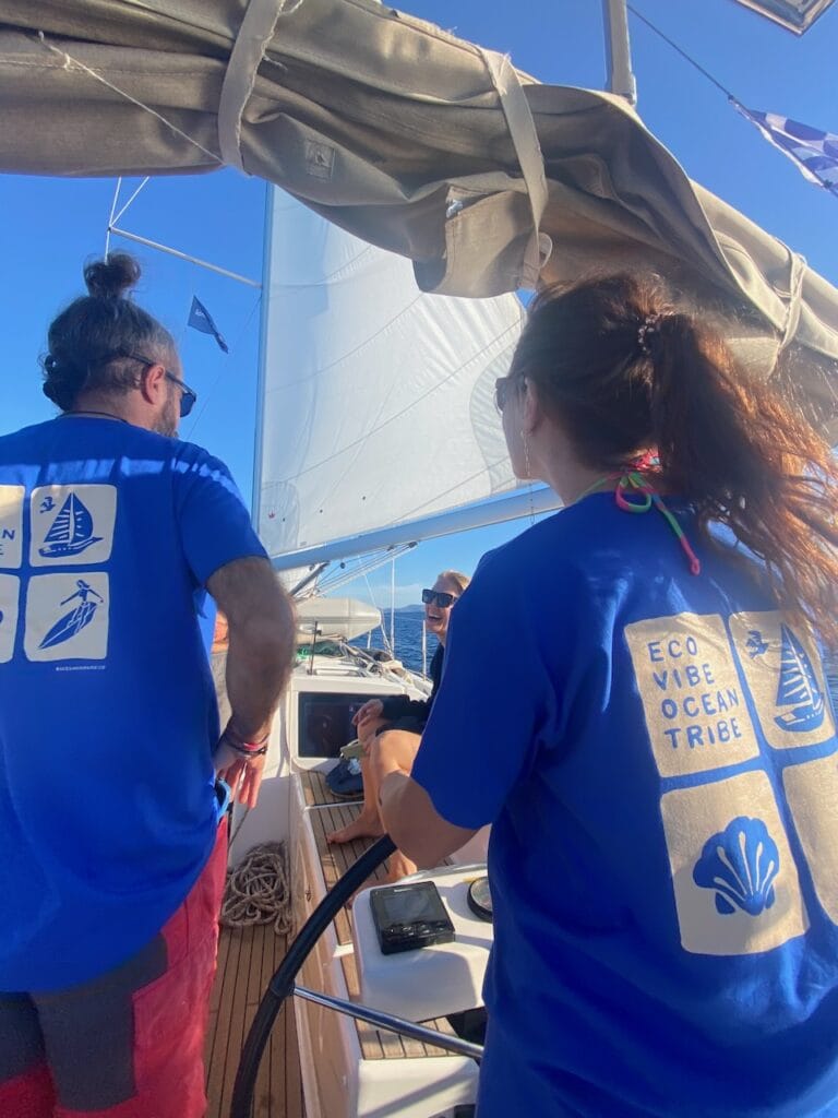 Two people wearing blue shirts with nautical designs are on a boat, engaged in sail training. One steers while the other stands beside them, enjoying the experience of offshore sailing near the Canary Islands. The sea and other boats paint a picturesque background.