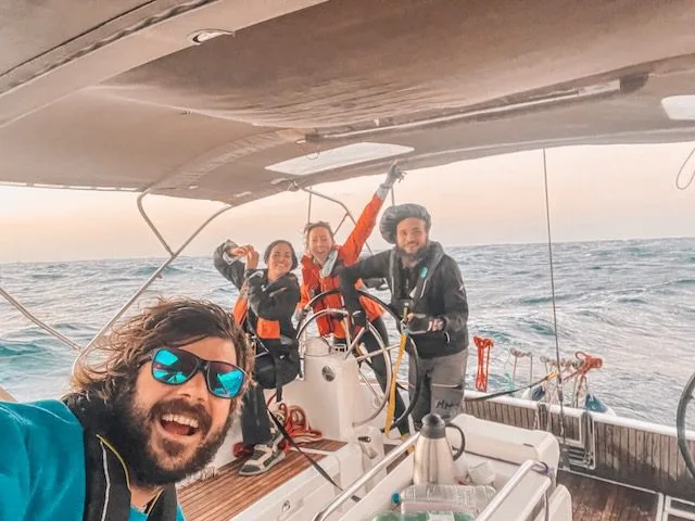 Four people smiling on a sailboat, with two steering and others standing around. The ocean, hinting at the adventure of sailing from the Canary Islands, is visible in the background under an overcast sky.