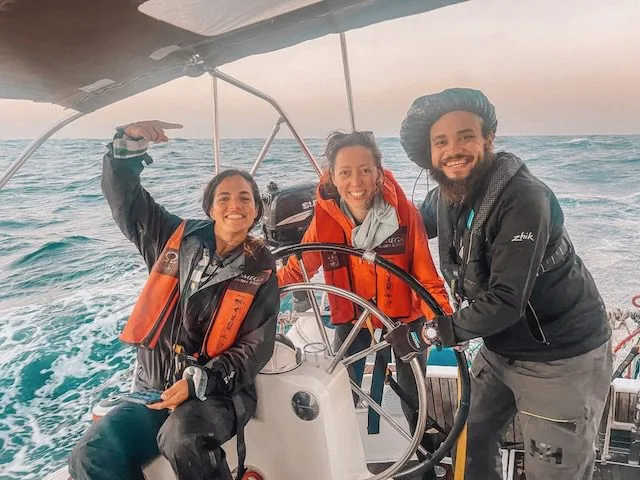 Three people on a sailing boat at sea, smiling and wearing life jackets, as they navigate the waters near Cape Verde. One person is confidently steering the wheel under the warm sun.