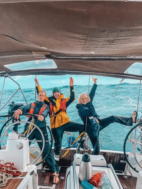 Three people on a boat, wearing jackets and life vests, pose playfully at the helm while sailing near the Canary Islands.