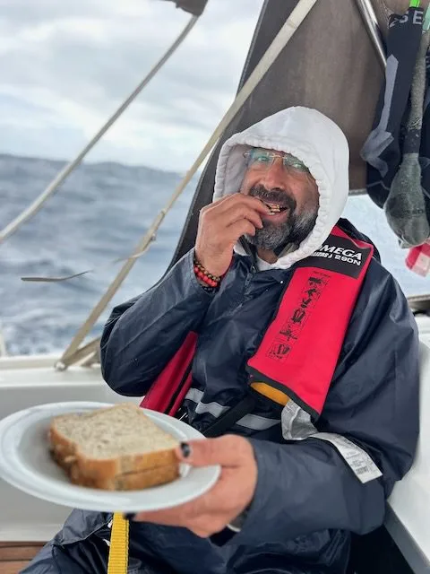 Person wearing a hooded rain jacket and life vest enjoys a sandwich aboard their boat, with the vast sea stretching out behind them. It's a perfect moment for sailing around the Canary Islands.