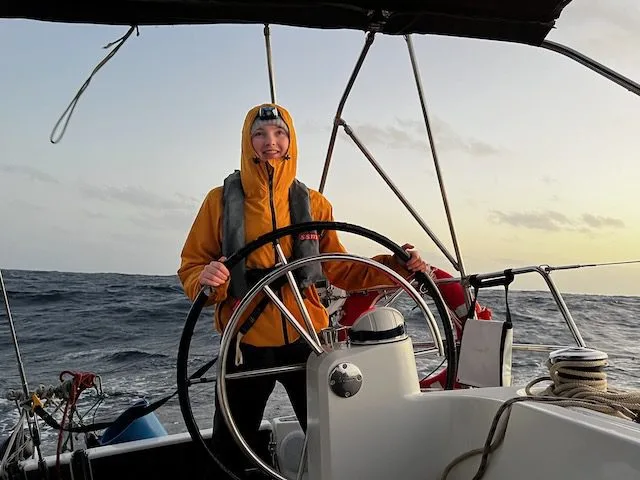 Person in a yellow jacket steering a sailboat at sea during sunset, with the stunning backdrop of Cape Verde on the horizon.