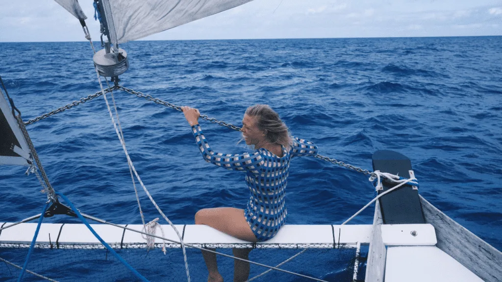 A woman in a patterned swimsuit sits on the edge of a sailboat, holding onto rigging as the wind sweeps over the open ocean beneath a cloudy sky—capturing a candid, documentary feel.