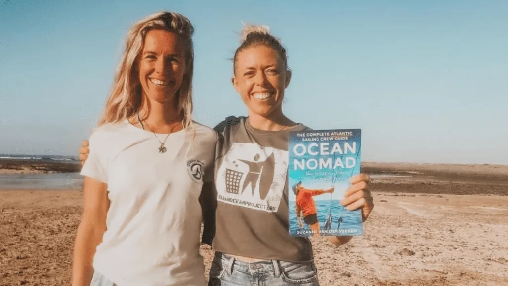 Two women stand together on a sandy beach, the wind gently tousling their hair as one holds a book titled "OCEAN NOMAD." Both are smiling and looking at the camera, with the sea and blue sky in the background.