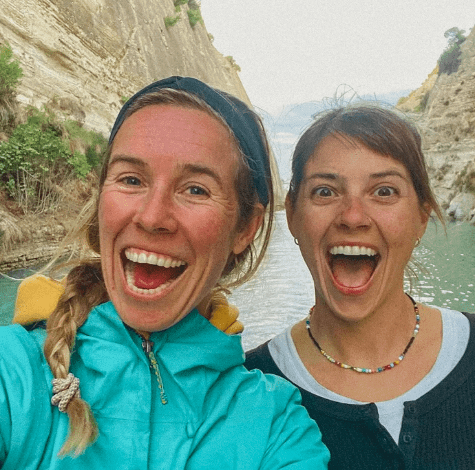 Two women stand outdoors in front of a rocky, water-filled canyon, smiling widely at the camera as they mark a special OCEAN NOMADS COMMUNITY celebration.