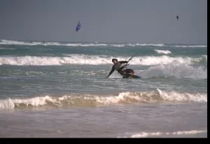 A person kite surfing near the shore, leaning back on the board with waves and another kite visible in the background.
